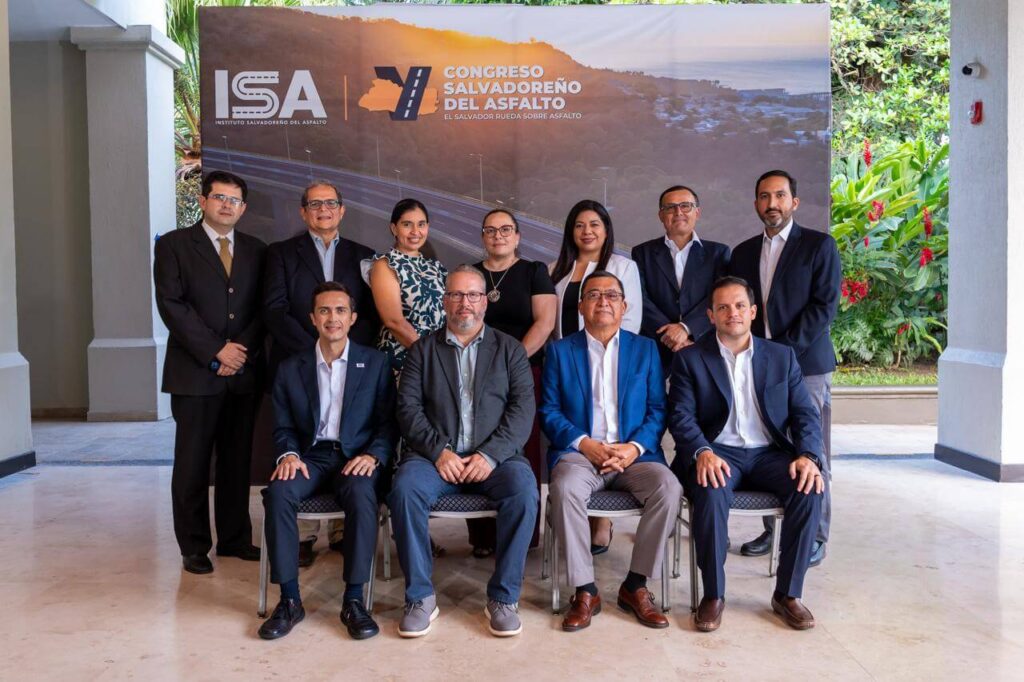 Diez personas en traje de negocios posan en dos filas delante de un telón de fondo en el que se lee "Congreso Salvadoreño del Asfalto" con vegetación de fondo.